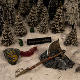 A 16cm metal Leviathan Axe replica resting on a dark rock in a snowy pine forest diorama, featuring a rock with Faye's yellow handprint and green ravens.
