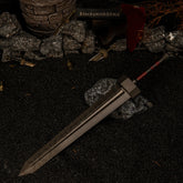 Greatsword metal replica resting diagonally on dark rocks, surrounded by black sand, scattered miniature broken swords, and small brass bells in a diorama setting.