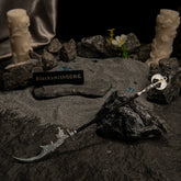 Loretta's War Sickle metal replica resting on dark rocks and grey sand, surrounded by white carved pillar candles, blue glintstone shards, and a white flower.