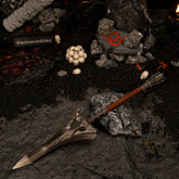 Serpent-Hunter metal replica resting on a dark volcanic rock, surrounded by black sand and miniature human skulls.