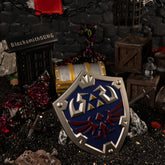 The complete 12cm metal Zelda Hylian Shield prop resting on dark gravel amidst a Hyrule Castle dungeon diorama, featuring a glowing Malice monster, a treasure chest, and an iron cage.