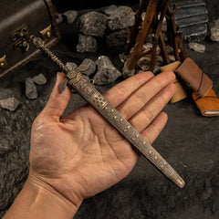 A hand holding the sheathed Banished Knight's Greatsword metal replica. The image shows the scale of the item relative to a human palm. The focus is on the detailed relief patterns on the bronze-colored scabbard. Background features leather journals and wood.