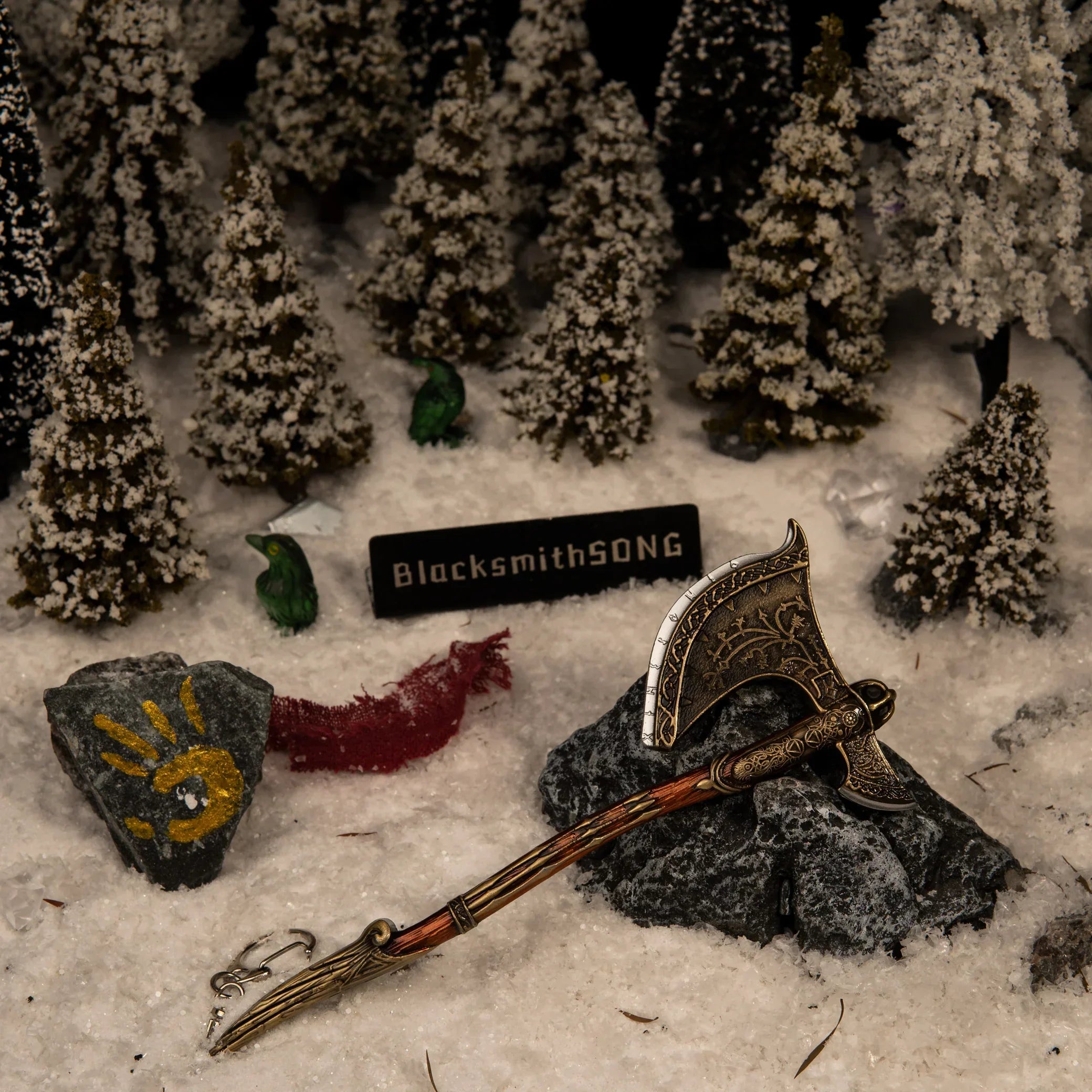 A 16cm metal Leviathan Axe replica resting on a dark rock in a snowy pine forest diorama, featuring a rock with Faye's yellow handprint and green ravens.