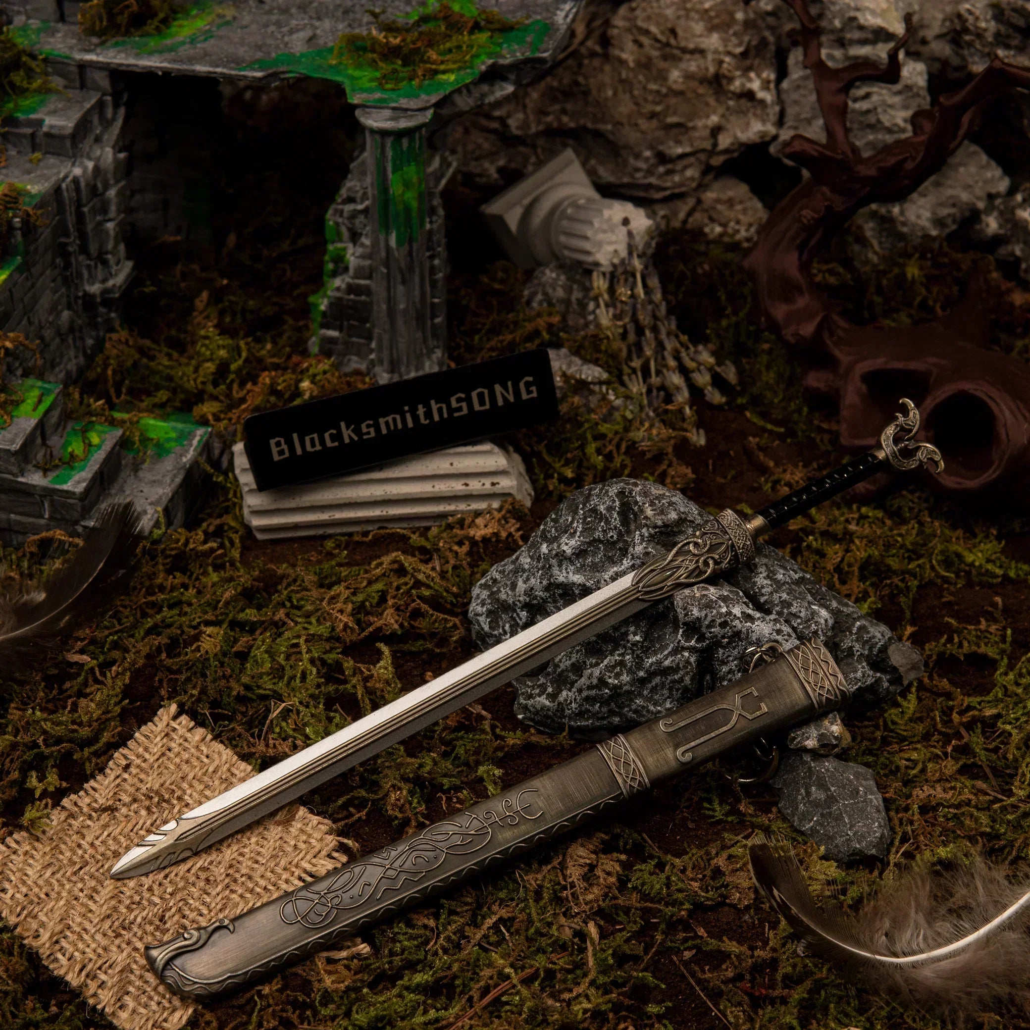 A 22cm silver metal Thrungva sword replica resting on a dark rock next to its highly detailed bronze-colored scabbard in a mossy ruins diorama.
