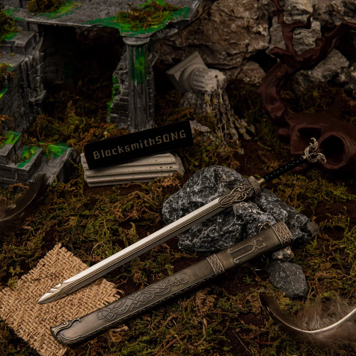 A 22cm silver metal Thrungva sword replica resting on a dark rock next to its highly detailed bronze-colored scabbard in a mossy ruins diorama.