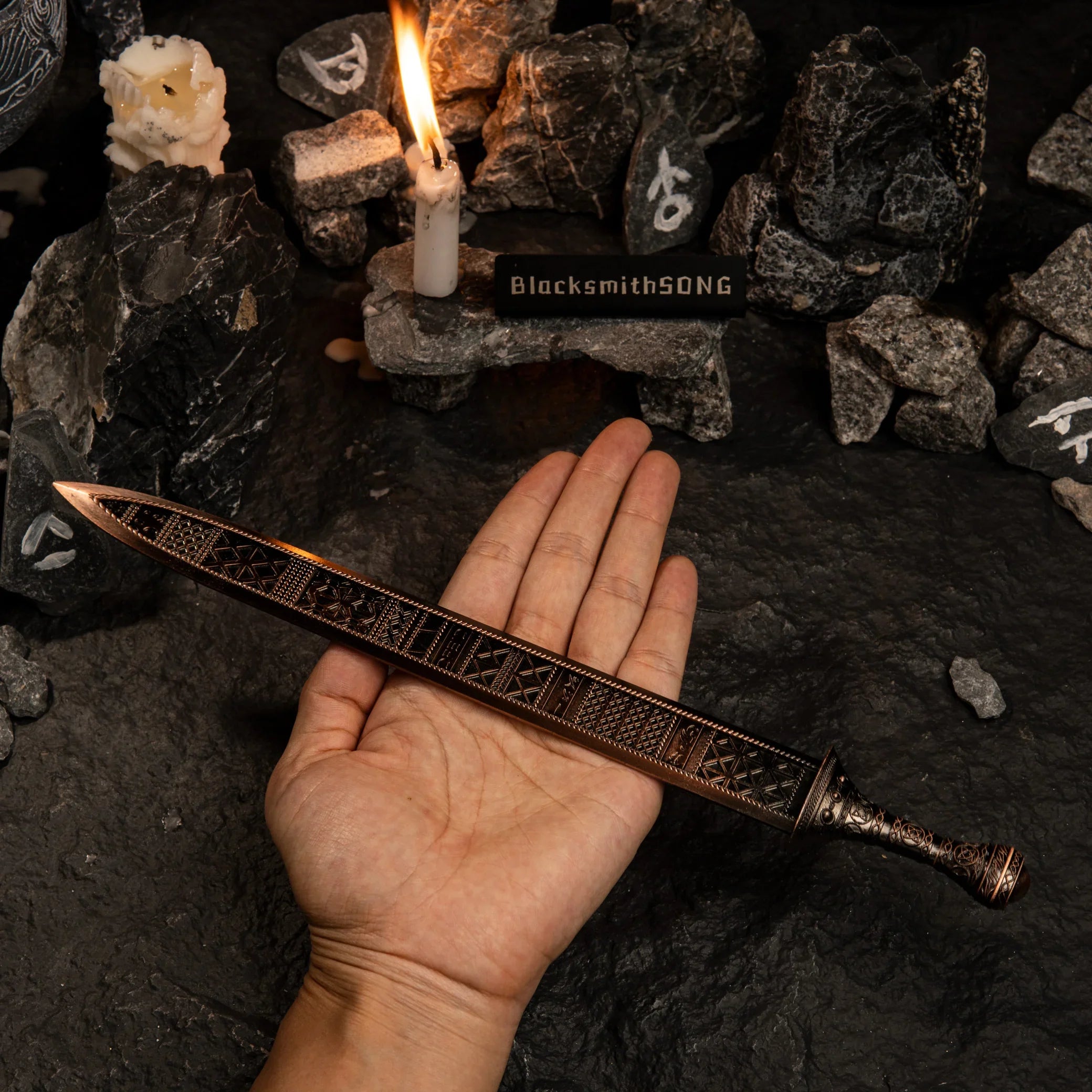 A hand holding the Regalia of Eochaid metal sword across the palm. The image shows the scale of the replica and highlights the intricate bronze-colored geometric designs on the blade surface. Background features dark rocks and a candle.