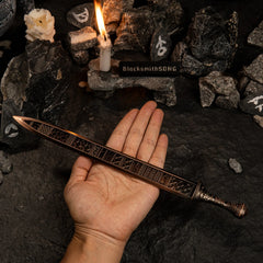 A hand holding the Regalia of Eochaid metal sword across the palm. The image shows the scale of the replica and highlights the intricate bronze-colored geometric designs on the blade surface. Background features dark rocks and a candle.