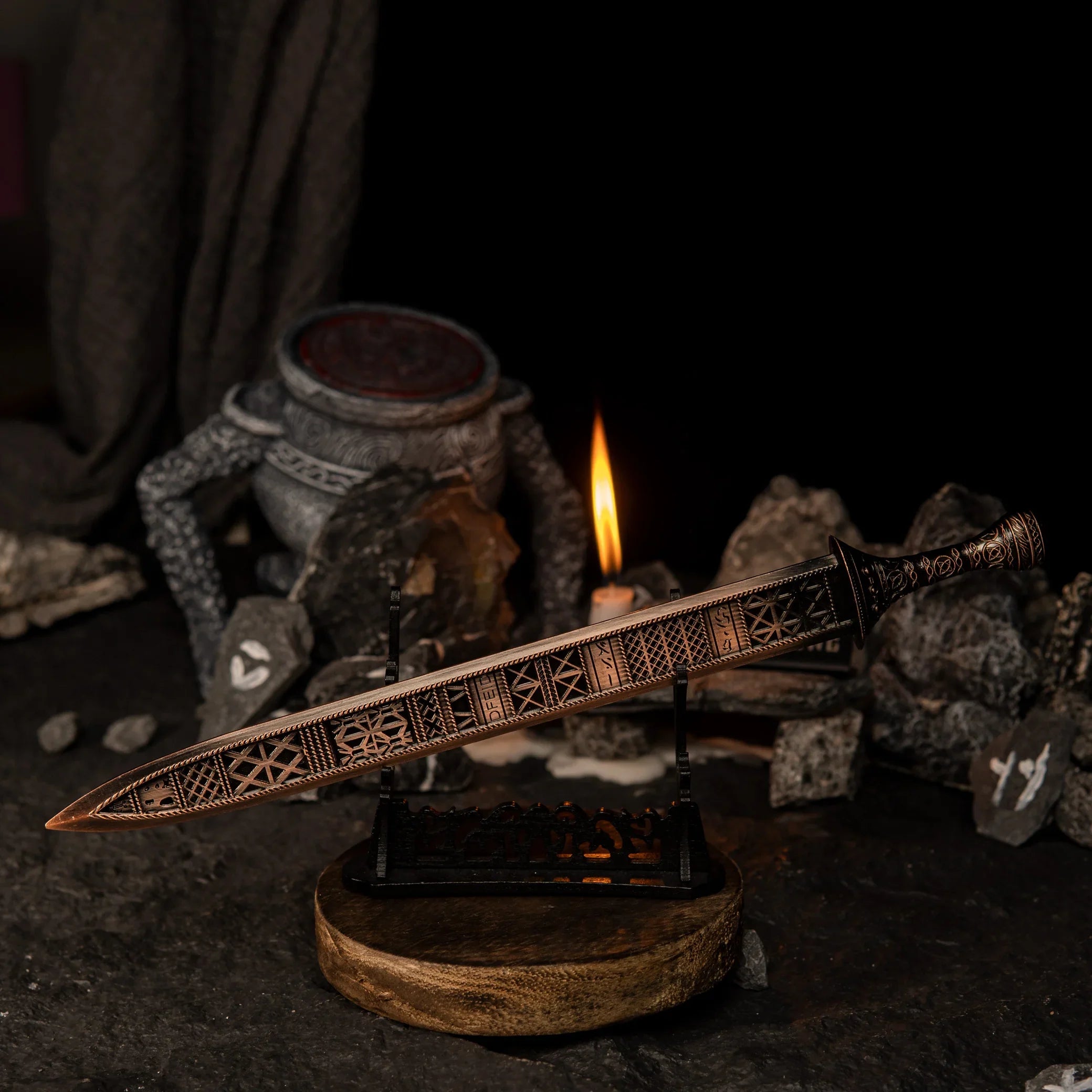 The Regalia of Eochaid sword replica from Elden Ring displayed on a black stand. The sword has a distinct bronze color with geometric patterns. It is surrounded by dark rocks painted with white rune symbols and a lit candle in the background. - Large display stand