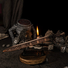 The Regalia of Eochaid sword replica from Elden Ring displayed on a black stand. The sword has a distinct bronze color with geometric patterns. It is surrounded by dark rocks painted with white rune symbols and a lit candle in the background. - Large display stand