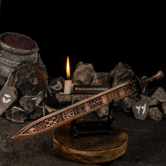 The Regalia of Eochaid sword replica from Elden Ring displayed on a black stand. The sword has a distinct bronze color with geometric patterns. It is surrounded by dark rocks painted with white rune symbols and a lit candle in the background. - Small display stand