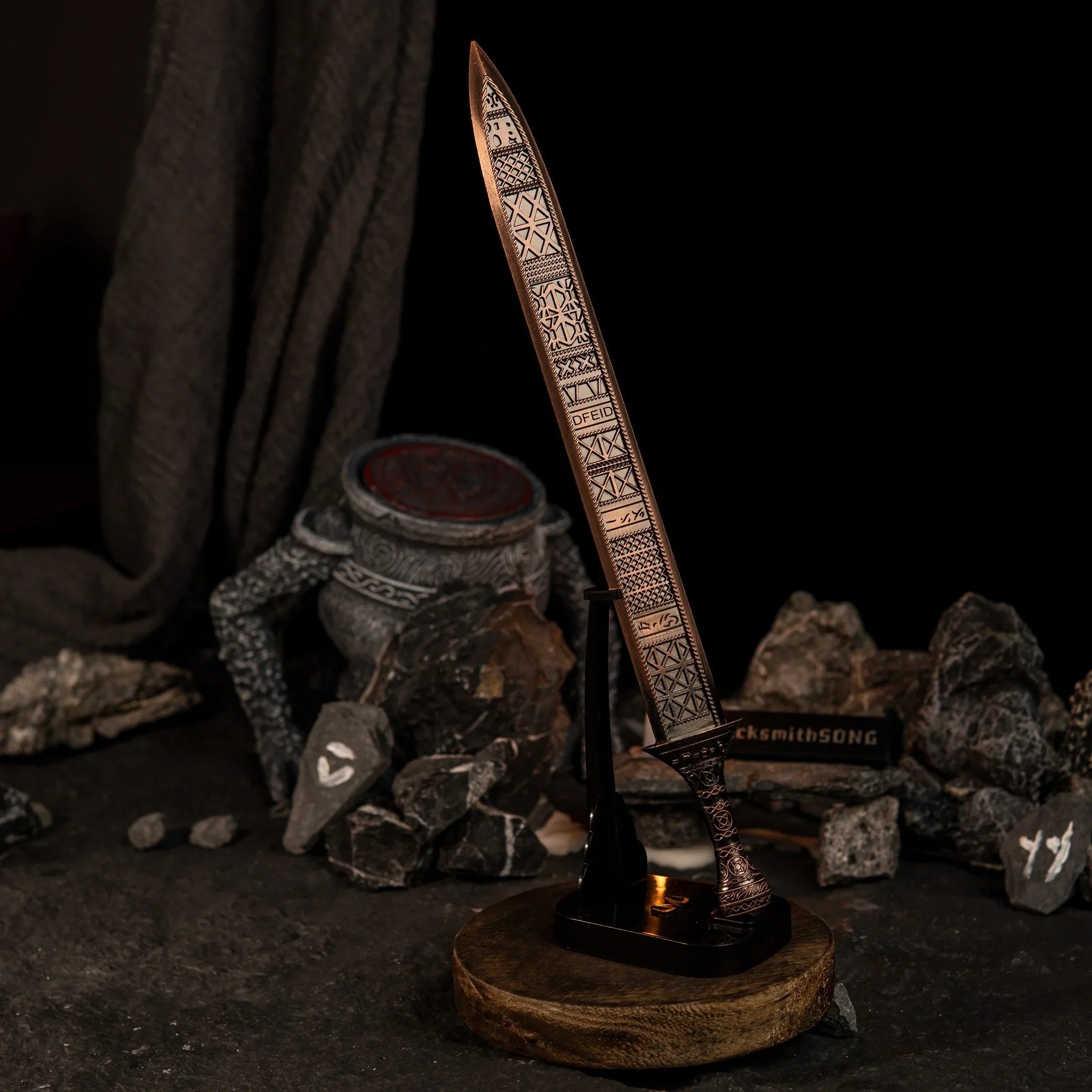 The Regalia of Eochaid sword replica from Elden Ring displayed on a black stand. The sword has a distinct bronze color with geometric patterns. It is surrounded by dark rocks painted with white rune symbols and a lit candle in the background. - Vertical display rack