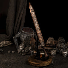 The Regalia of Eochaid sword replica from Elden Ring displayed on a black stand. The sword has a distinct bronze color with geometric patterns. It is surrounded by dark rocks painted with white rune symbols and a lit candle in the background. - Vertical display rack