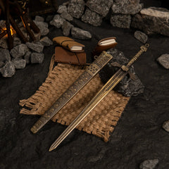 Banished Knight's Greatsword replica resting on burlap and dark rocks. Background features a miniature campfire and small leather journals.