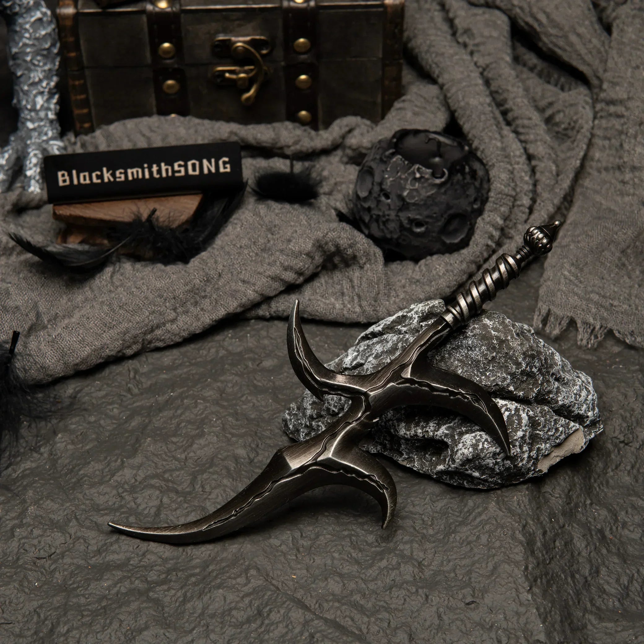 Black Knife metal replica resting on rocks. Background features black feathers, grey fabric, and a moon-like spherical prop.