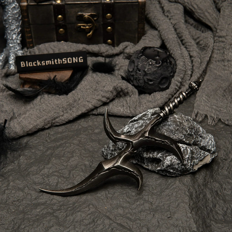 Black Knife metal replica resting on rocks. Background features black feathers, grey fabric, and a moon-like spherical prop.