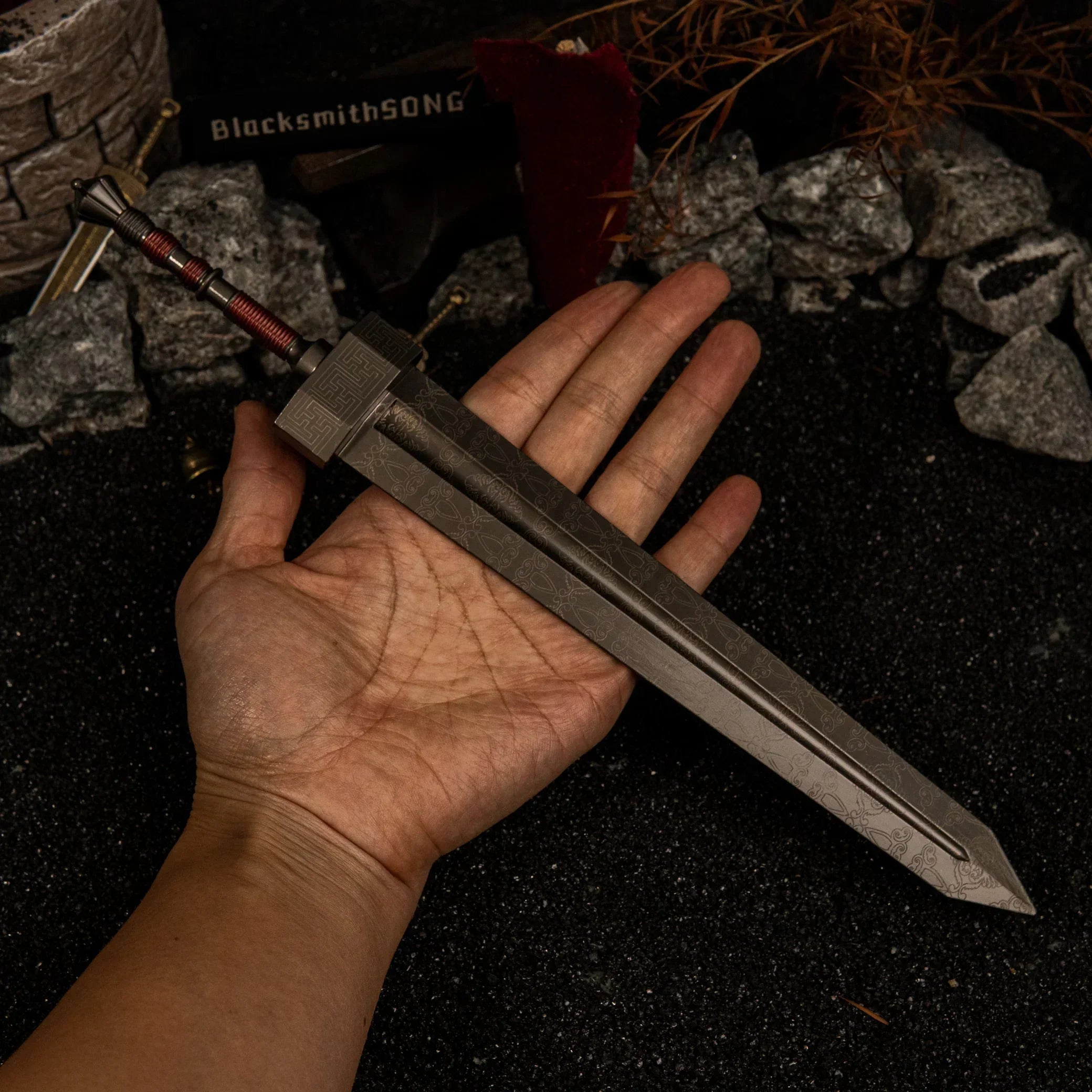 A hand holding the massive 30cm Greatsword metal replica to show its scale, highlighting the wide grey blade and red-wrapped handle against a dark background.