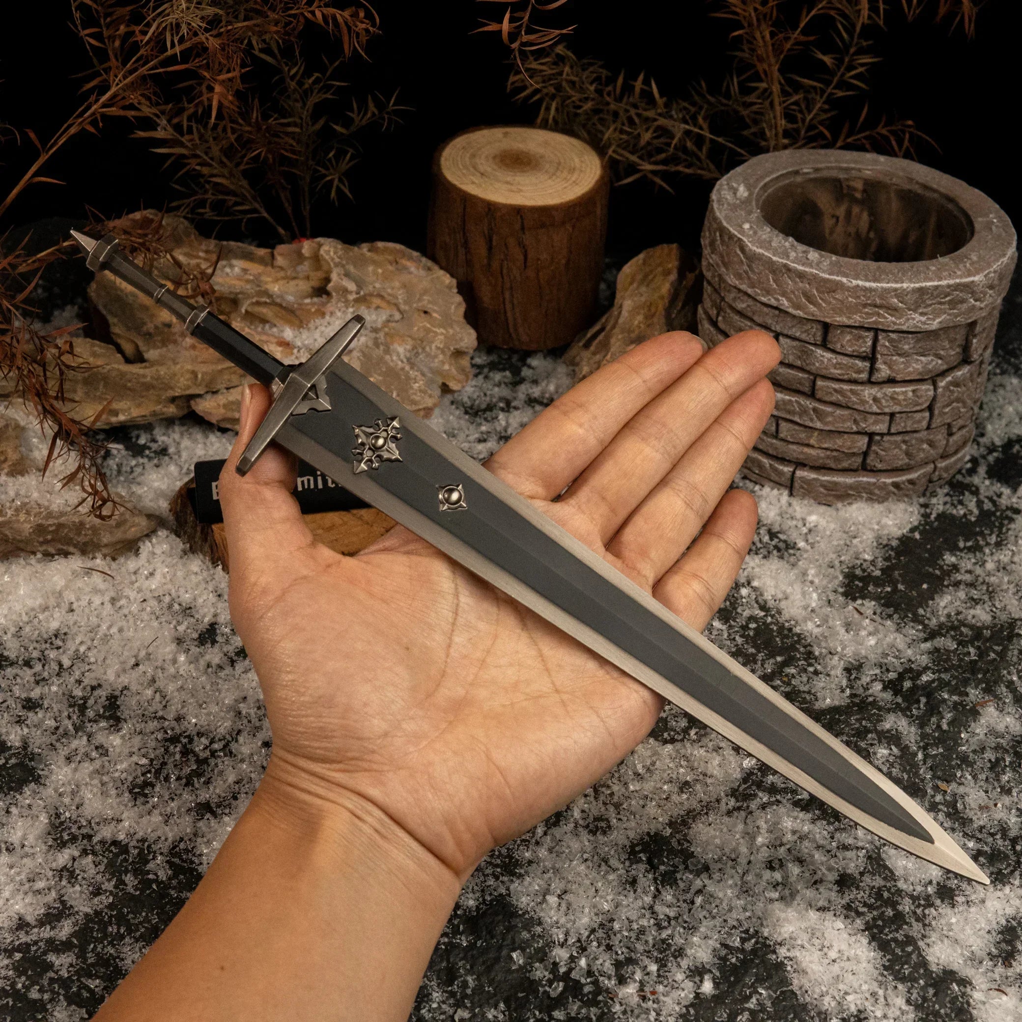 A hand holding the Greatsword of Solitude replica to show its scale, resting over a background of snow, rocks, and dry foliage.