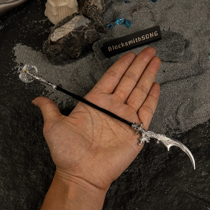 A hand holding the 22cm Loretta's War Sickle metal replica to demonstrate its long scale, set against a dark grey sand background.