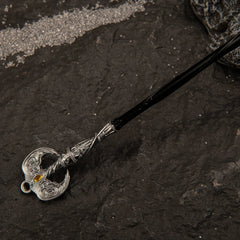 Detailed view of the ornate silver pommel of the Loretta's War Sickle miniature, featuring wing-like metal carvings and a yellow gem accent.