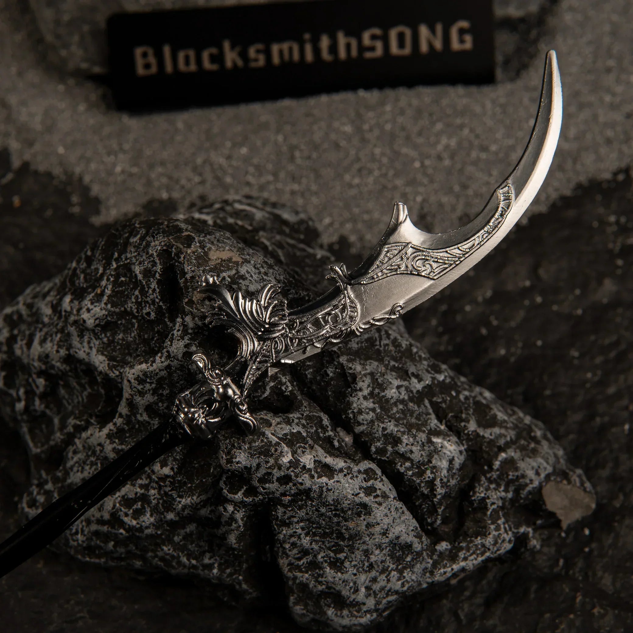 Macro close-up shot of the silver crescent blade of Loretta's War Sickle replica, showcasing intricate floral engravings resting on a dark rock.