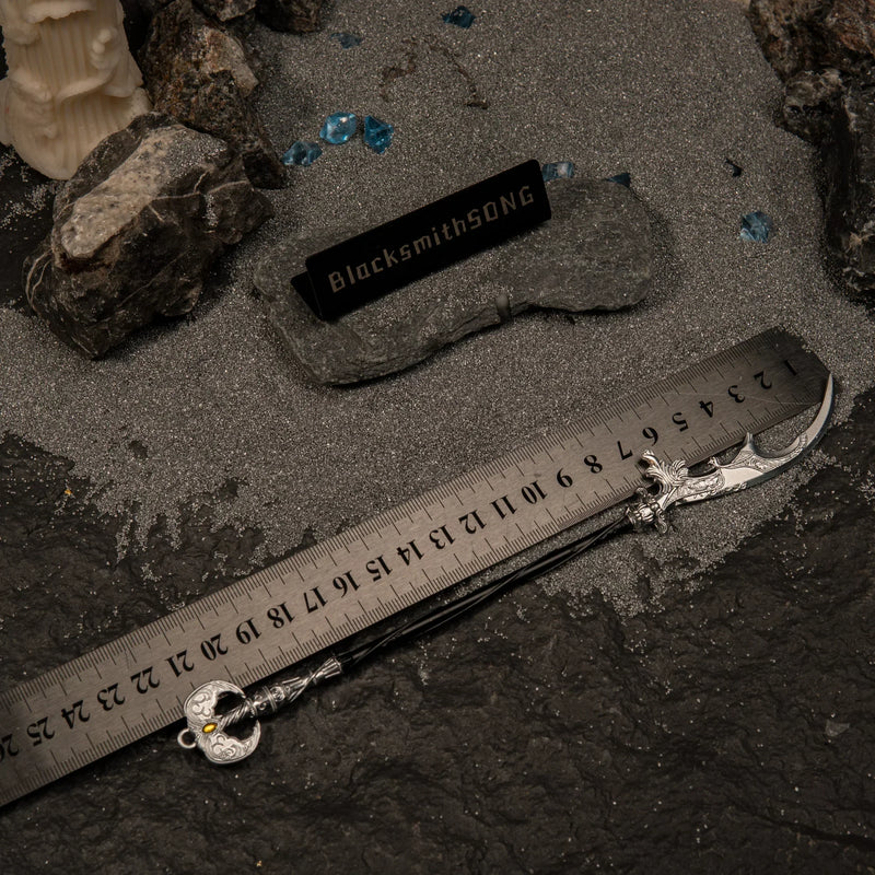 Loretta's War Sickle metal replica placed next to a measuring ruler on grey sand, indicating a total length of approximately 22 centimeters.