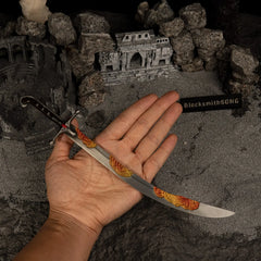 A hand holding the 26cm Nomad's Kilij metal replica to show its scale, highlighting the curved blade and frenzied flame colored engravings against grey sand.