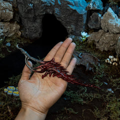 Human hand holding the 22cm Reduvia replica, demonstrating its size, 98g heavy weight, and crimson blood-coated finish.
