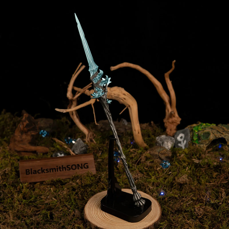 Vertical display of the Spear of the Dreaming replica on an upright black stand, set against a dark background with green moss and driftwood branches.