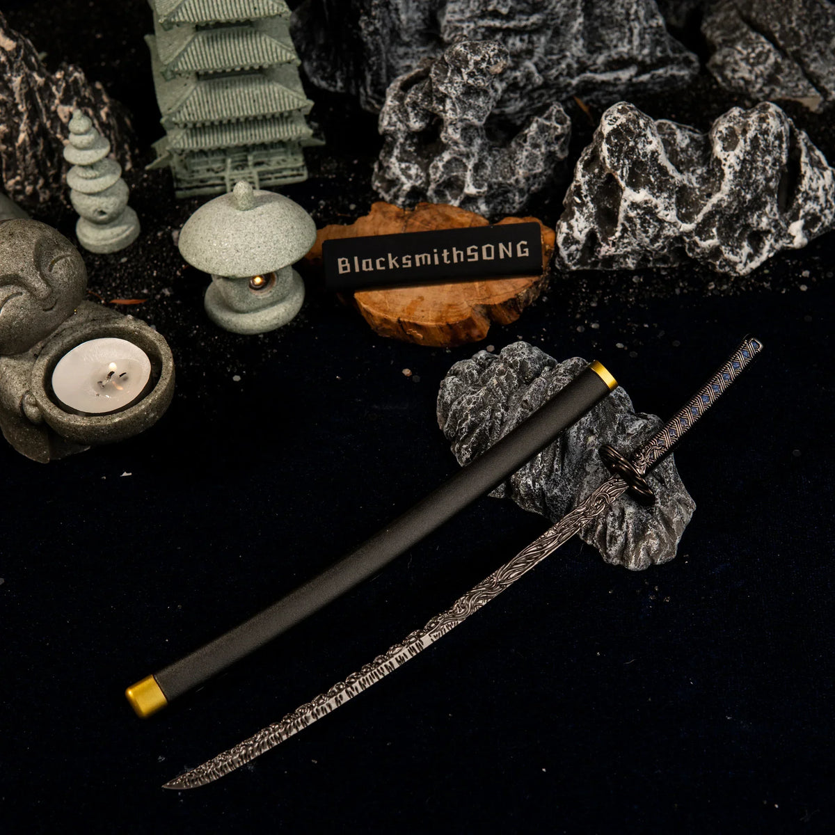 Star Shadow metal katana and scabbard resting on dark rocks in a Zen garden diorama, featuring a miniature stone pagoda, lantern, and Buddha statue.