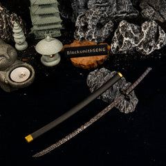 Star Shadow metal katana and scabbard resting on dark rocks in a Zen garden diorama, featuring a miniature stone pagoda, lantern, and Buddha statue.