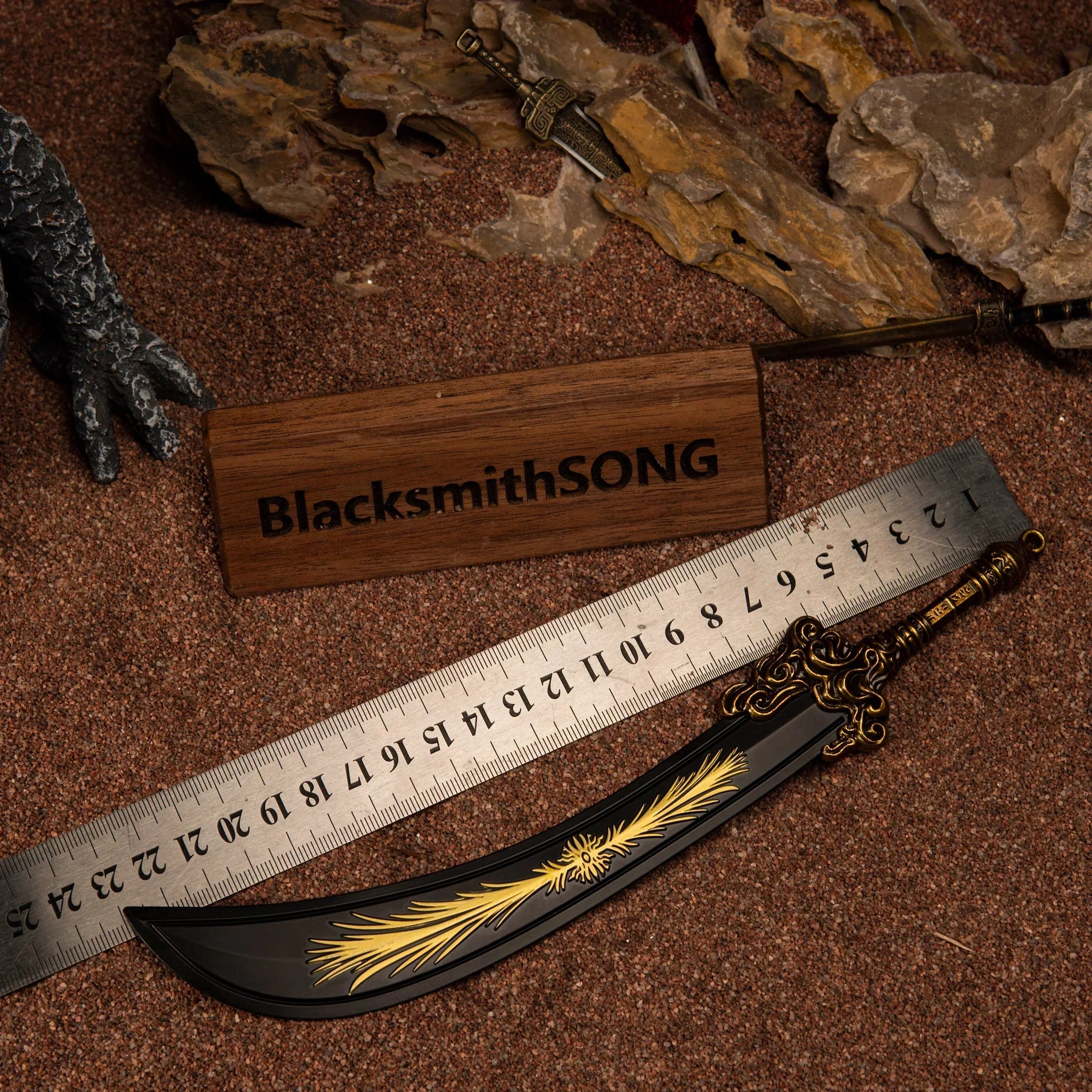 A Starscourge Greatsword metal replica placed next to a measuring ruler on red sand, indicating a length of approximately 23 centimeters.