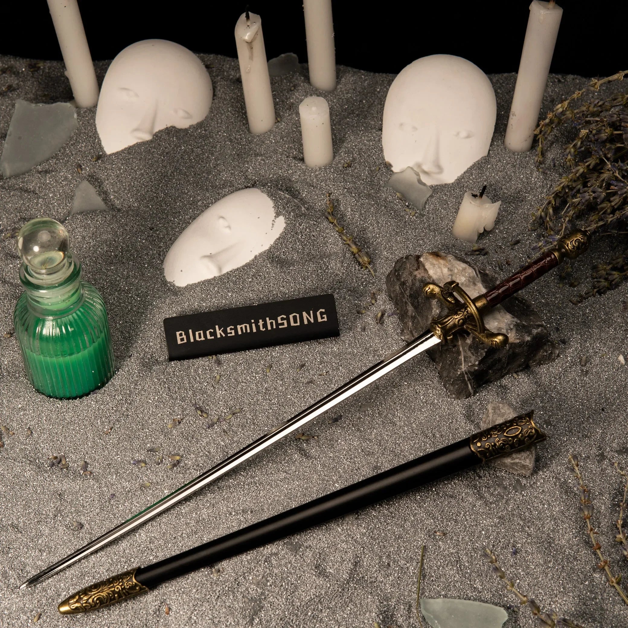 Needle metal sword replica unsheathed on grey sand, surrounded by white masks, candles, and a vial of poison.