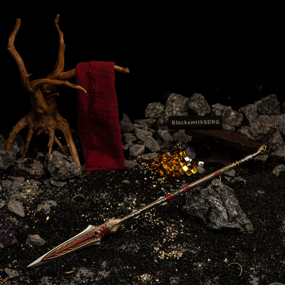 A 35cm metal Draupnir Spear replica resting on dark volcanic sand, with a blacksmith anvil, golden rings, and a red Spartan cloth on a dead tree in the background.
