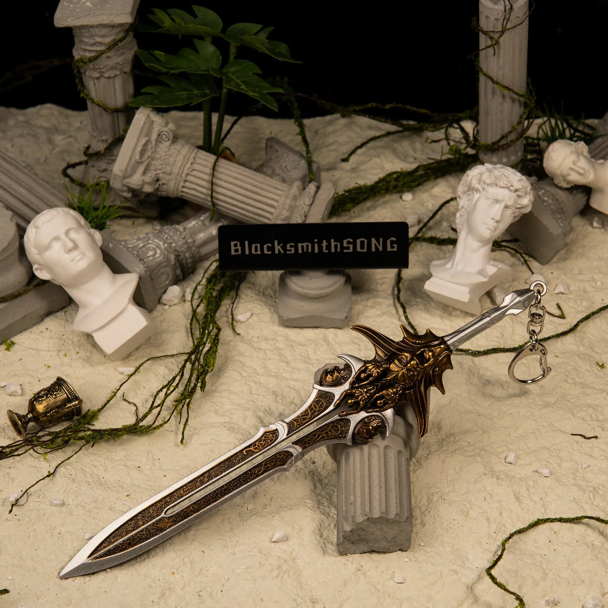 A 21cm metal Blade of Olympus replica resting on a broken Greek column surrounded by white sand, shattered marble busts, and green vines.