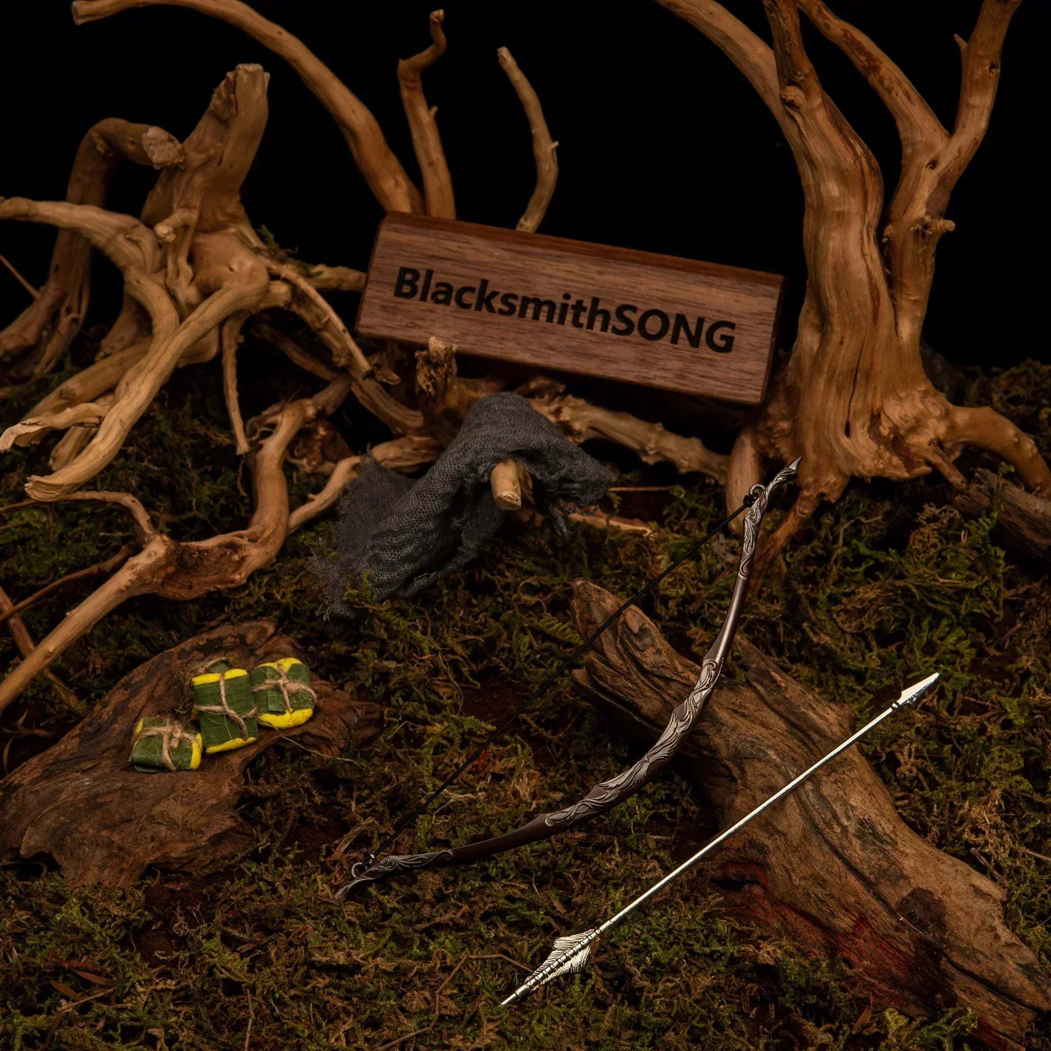 Bow of the Galadhrim metal replica and arrow resting on green moss, surrounded by twisted roots, a grey cloak, and wrapped Lembas bread.
