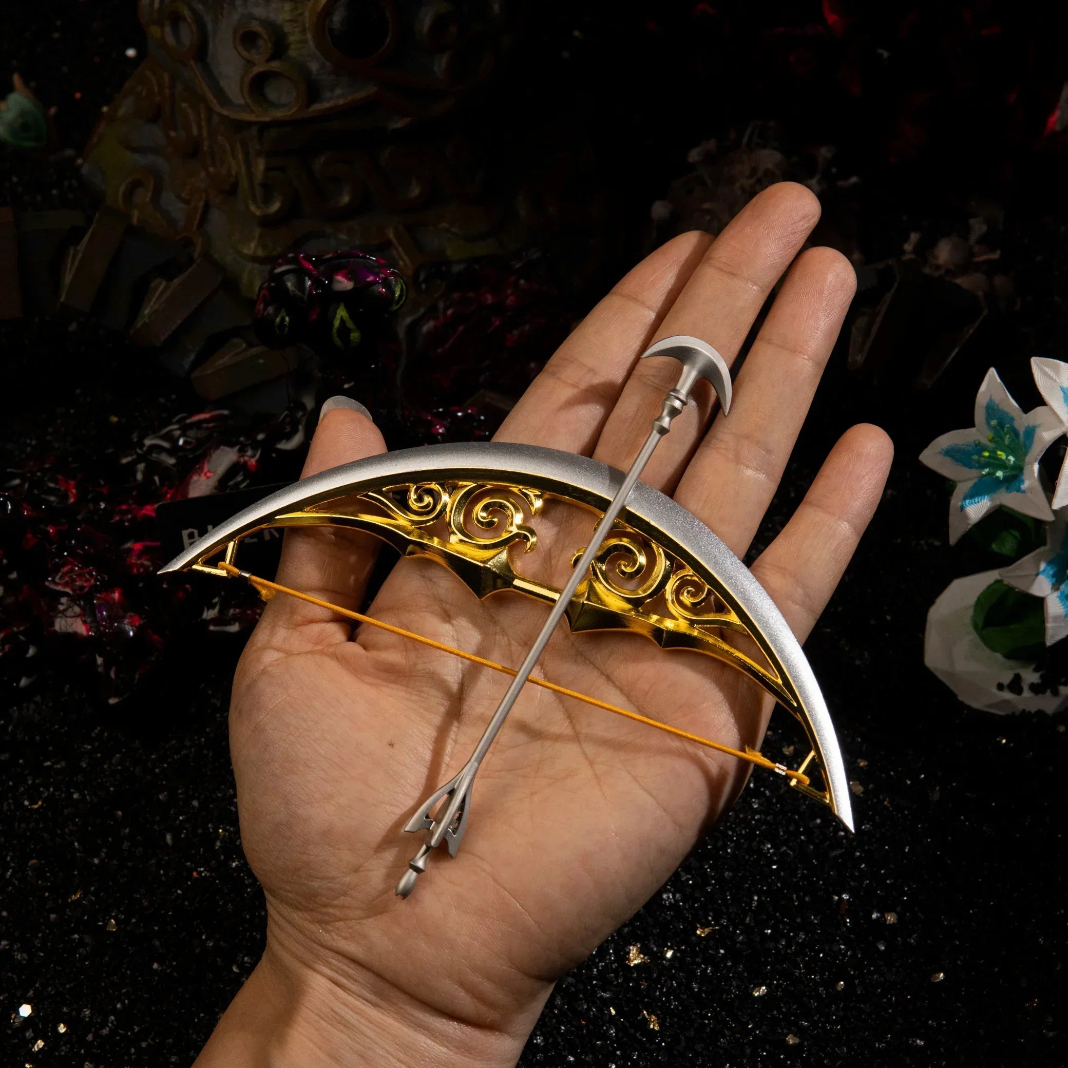 An adult hand displaying the 74g heavy metal Bow of Light miniature and the separate Light Arrow, demonstrating the 15cm scale and the elastic bowstring.