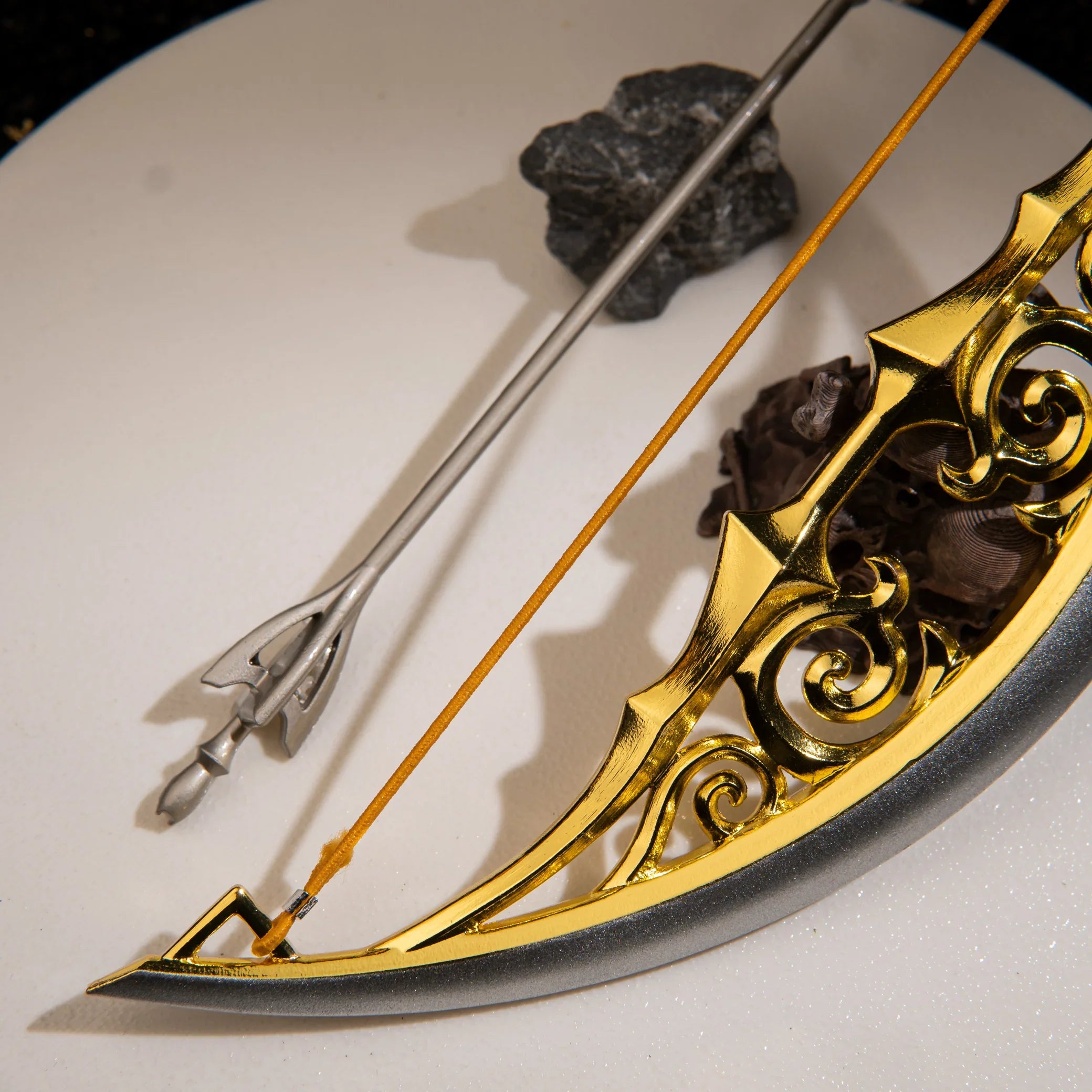 Detailed view of the 12.5cm silver Light Arrow and the taut elastic string of the metal Bow of Light miniature, resting on a white pedestal.