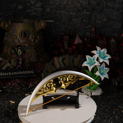 The metal Bow of Light replica resting symmetrically on a compact black desk stand, with the silver Light Arrow lying across the white base amidst dark crystals and blue flowers.