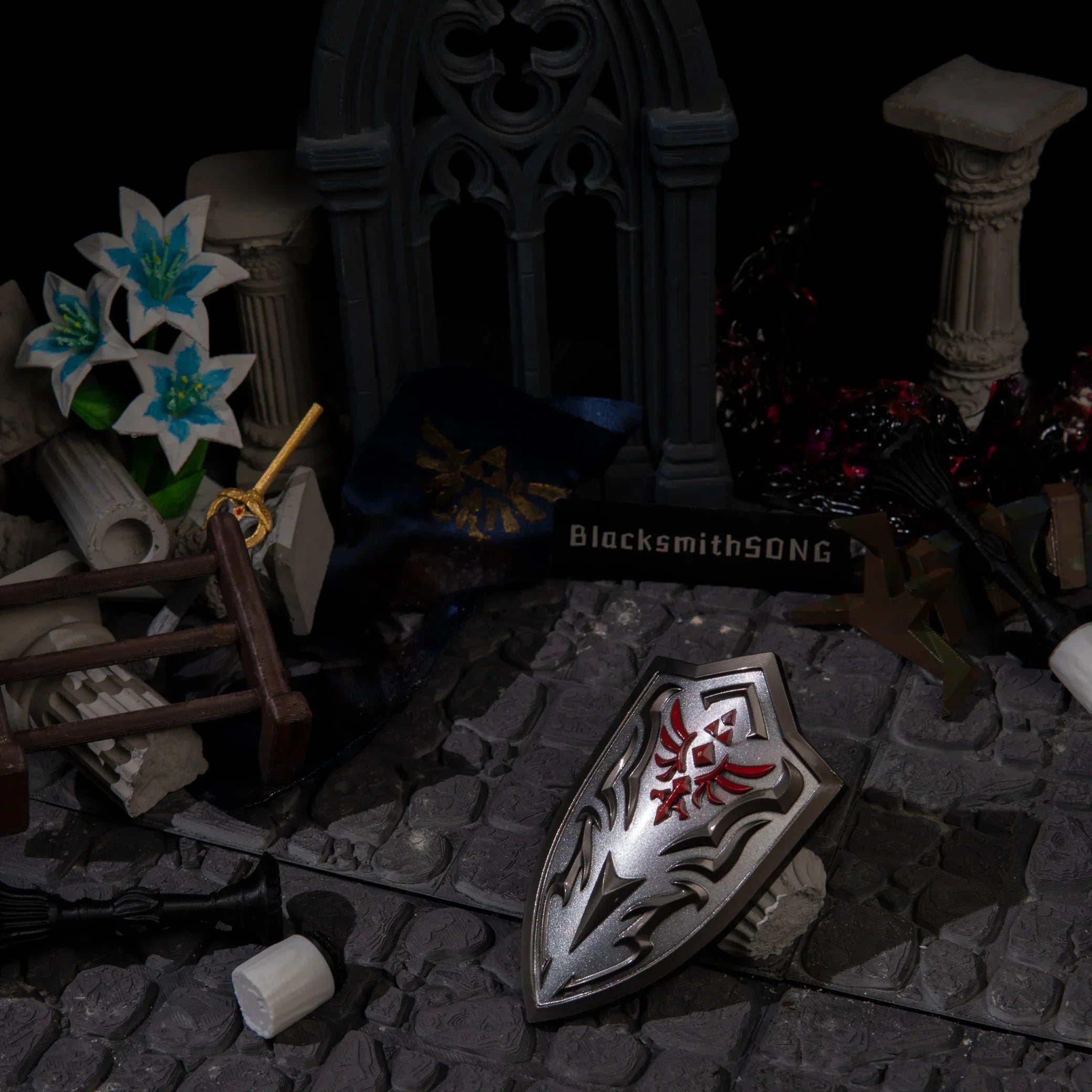Royal Guard's Shield metal miniature displayed on cobblestones, surrounded by Ganon's Malice and Silent Princess flowers.