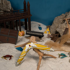 Sea-Breeze Boomerang replica (14cm die-cast zinc alloy) — propped on driftwood in a handmade beach diorama, by BlacksmithSONG.