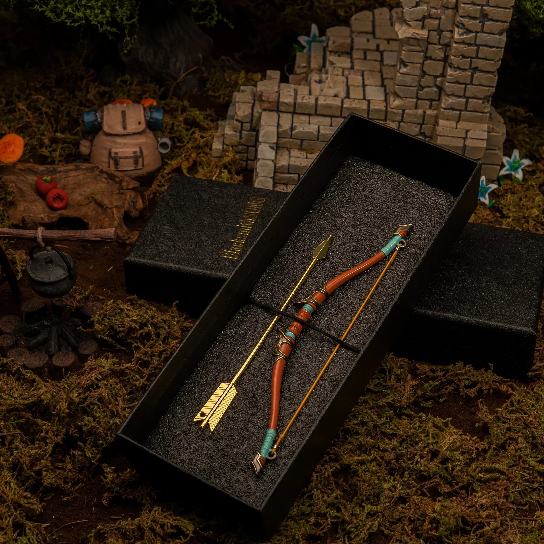 1/6 scale Traveler's Bow metal miniature and golden arrow inside a black gift box, placed near a Korok Backpack diorama prop.