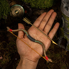 An adult hand displaying the 52g heavy metal Zelda Wooden Bow miniature and the separate golden arrow, demonstrating the 18cm scale and elastic string.