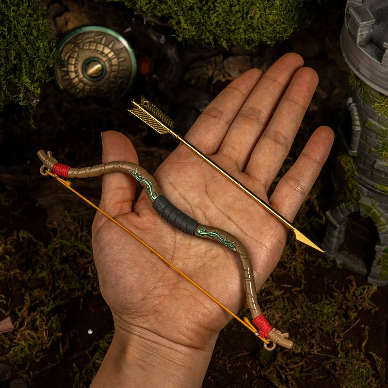 An adult hand displaying the 52g heavy metal Zelda Wooden Bow miniature and the separate golden arrow, demonstrating the 18cm scale and elastic string.
