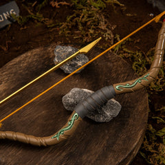 Close-up shot of the Zelda Wooden Bow replica's center grip, highlighting the sculpted dark leather texture, green vine motifs, and the nocked golden arrow resting on a wooden slice.
